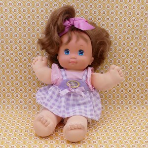Puede incluir: Un vestido de cuadros vichy morado y blanco con un lazo rosa en el pelo de una muñeca con pelo castaño y ojos azules.