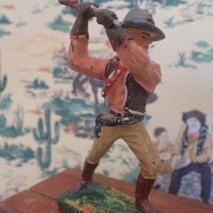 May include: A toy cowboy figurine with a brown hat, pink shirt, and brown waistcoat, holding a rifle. The figurine is standing on a green base.