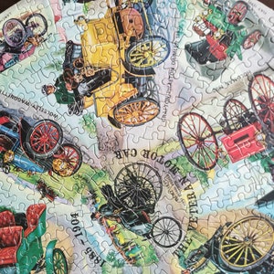 May include: A vintage jigsaw puzzle depicting a collection of antique automobiles from the late 19th and early 20th centuries. The puzzle features various makes and models, including a Wolseley Wagonette, a Lanchester, a Benz, a Craphton Phaeton Daimler, and a Holgman. The puzzle pieces are arranged in a circular pattern, with the names of the cars and their years of manufacture printed on the puzzle pieces.