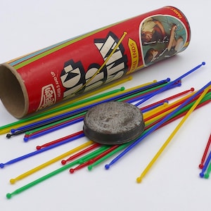Può includere: Un gioco vintage di bastoncini in un tubo rosso e giallo con le parole "Pick Up Sticks" e "Small Tall Ends" stampate sul lato. Il gioco include una varietà di bastoncini colorati e una base in metallo.