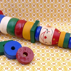 Puede incluir: Un juguete de madera colorido con una cara de payaso y varias formas geométricas. El juguete presenta elementos pintados en rojo, azul, verde, blanco y amarillo. Las formas sueltas adicionales incluyen un cuadrado verde y círculos rojos y azules.