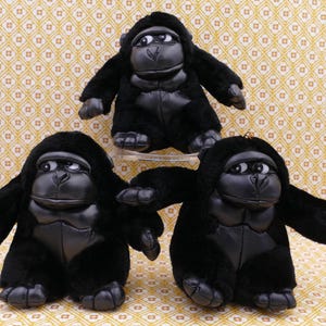 May include: Three black plush gorilla toys with leather-look faces, hands, and feet. The gorillas have large eyes and are in a sitting position. The background is a patterned yellow and white fabric.