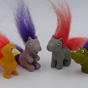 May include: Four small plush toys with colourful hair. A yellow bird with a pink tail, a grey horse with a purple tail, a grey horse with a pink tail, and a green alligator with a purple tail.