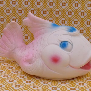 May include: A vintage-style, white fish-shaped toy with pink and blue details. The toy features a smiling face with blue eyes, pink cheeks, and an open mouth. The fish is set against a yellow and white patterned background.