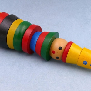 Puede incluir: Un juguete de madera colorido con forma de oruga. El juguete presenta una serie de anillos de madera de colores brillantes en amarillo, rojo, negro, verde y azul. La cabeza es de color canela claro con ojos negros y nariz roja. Fondo azul claro.