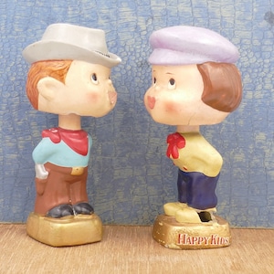 May include: Two vintage bobblehead figurines. One figure wears a cowboy hat, blue shirt, red scarf, and brown pants. The other figure wears a purple cap, yellow shirt, red bow tie, and blue pants. The bases are gold with "HAPPY KIDS" text.