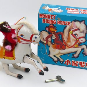 May include: A white wind-up toy horse with a red saddle and a brown monkey rider. The horse has a white mane and tail. The toy is in its original box, which is blue with white stars and the words "Monkey Riding Horse".