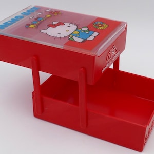 Puede incluir: Una caja de notas de plástico rojo con una tapa transparente con un diseño de Hello Kitty. La caja tiene un cajón extraíble y un asa. La tapa tiene la inscripción "MEMO BOX".