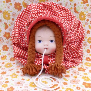 May include: A vintage doll with blue eyes and brown yarn braids, wearing a red and white patterned bonnet. A white string extends from the doll's mouth. The background features a floral pattern.