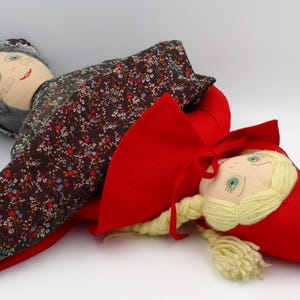 Puede incluir: Dos muñecas de tela hechas a mano. Una representa a una abuela con un vestido floral y pelo gris, la otra una niña con una capa roja. Las muñecas tienen caras pintadas y están tumbadas.