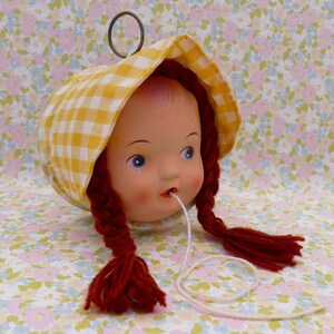 May include: A vintage doll head with blue eyes, red yarn braids, and a yellow and white checkered bonnet. A metal ring is attached to the top of the head. A white string extends from the doll's mouth. The doll is set against a floral background.