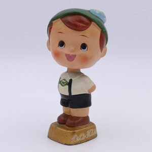 May include: A vintage figurine of a smiling boy wearing a green hat with a blue pom-pom, a white shirt with a green emblem, black shorts, and brown shoes. The figurine stands on a gold-colored base with the text "Let's Kiss".