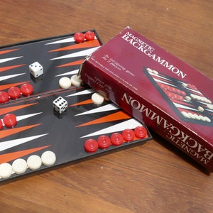 Puede incluir: Un juego de backgammon magnético rojo y negro con fichas blancas y rojas. El juego está abierto y listo para jugar. La caja es roja con letras blancas y muestra una imagen del juego.