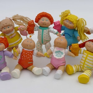 May include: A group of eight Cabbage Patch Kids dolls, each with different colored clothing and accessories. The dolls are all sitting or standing on a white surface.