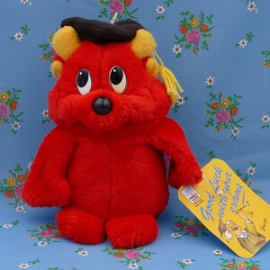 Pode incluir: Urso de pelúcia vermelho usando um chapéu de formatura preto com uma borla amarela. O urso tem olhos grandes e pretos e um nariz preto. O urso está segurando uma etiqueta amarela que diz "Boa sorte com seus sonhos".
