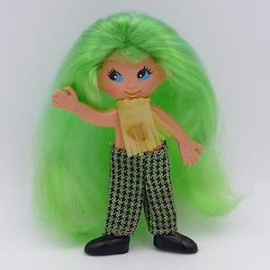 May include: A doll with green hair and blue eyes, wearing a yellow fringed top and black and white checked trousers.