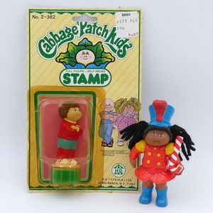 Może przedstawiać: Zestaw vintage stempla i lalki Cabbage Patch Kids. Stempel to pełnopostaciowy, samonaprawiający się wzór. Lalka ubrana jest w czerwony mundur z niebieskim kapeluszem i butami, trzymając laskę cukrową.