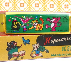 May include: Two vintage harmonicas, one atop the other, with colorful illustrations. The top harmonica has a green background with cartoon animals. The bottom box reads "Harmonica" and "UC 513" with more cartoon characters.