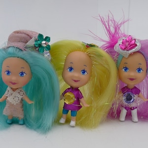 Könnte beinhalten: Drei kleine Puppen mit langen, bunten Haaren. Die Puppe links hat türkisfarbenes Haar und einen rosa-grünen Hut. Die Puppe in der Mitte hat gelbes Haar und ein lilafarbenes Shirt. Die Puppe rechts hat pinkfarbenes Haar und eine silber-rosa Krone.