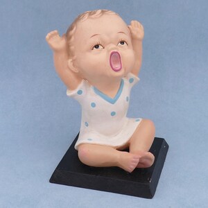 May include: A ceramic figurine of a baby with its arms raised, wearing a white dress with blue polka dots and a blue collar. The baby is sitting on a black base.