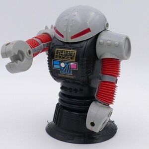 May include: A vintage Robo Force toy robot, primarily black with gray and red accents. The robot has a helmet-like head, articulated arms, and the Robo Force logo on its chest. The text "HUNDRED" is also visible.
