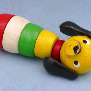 Puede incluir: Un juguete de madera colorido con forma de perro, con un cuerpo segmentado y una cabeza redonda amarilla. El cuerpo está hecho de anillos de madera rojos, blancos, verdes y amarillos. El perro tiene orejas negras y ojos blancos.