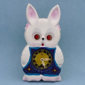 May include: White ceramic bunny clock with red eyes and a blue and yellow clock face. The clock face has black numbers and hands.