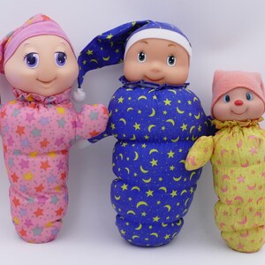 Puede incluir: Tres muñecos de peluche con caras suaves y redondeadas y gorros de dormir. El muñeco de la izquierda es rosa con estrellas coloridas, el central es azul con lunas y estrellas amarillas, y el de la derecha es amarillo con lunas y estrellas rosas.