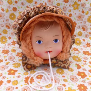 May include: A vintage doll head with blue eyes and rosy cheeks, wearing a brown and orange patterned bonnet and a knitted orange scarf. A white string extends from the doll's mouth. The background is a floral pattern.