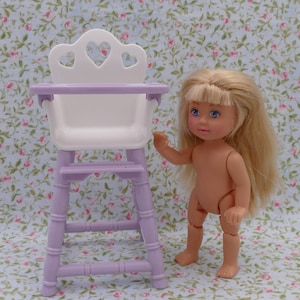 Puede incluir: Una silla alta para muñecas de color morado y blanco con un asiento blanco y un marco morado. La silla tiene tres recortes en forma de corazón en la parte posterior. Una muñeca rubia está de pie junto a la silla.