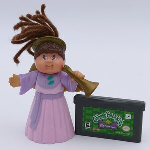 Puede incluir: Una figura de ángel de Cabbage Patch Kids con un vestido lavanda y una trompeta dorada, junto a un cartucho de videojuego Cabbage Patch Kids: The Patch Puppy Rescue. La figura tiene cabello de hilo marrón y una cara sonriente.