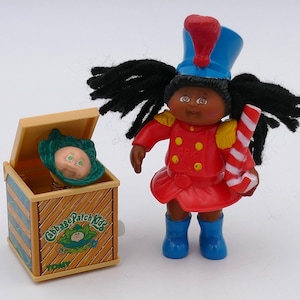 May include: A Cabbage Patch Kids toy set featuring a doll in a red uniform with a blue hat and boots, holding a candy cane. Beside it is a wooden box with a cabbage patch kid head popping out. The box has the Cabbage Patch Kids logo.