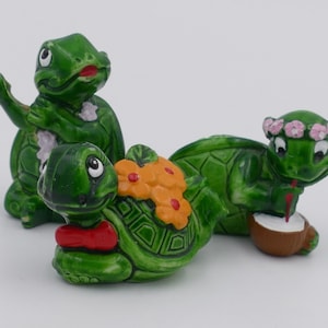 Puede incluir: Tres figuritas de tortugas de cerámica verde. Una tortuga lleva un collar de flores y sostiene un coco con una pajita. Otra tortuga lleva un collar de flores y sostiene una flor roja. La tercera tortuga sostiene una flor roja.