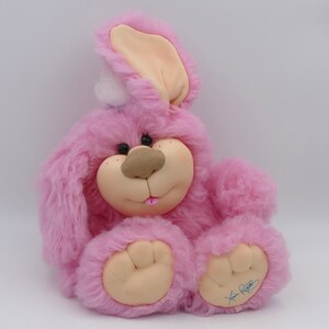 Puede incluir: Conejo de peluche rosa con un pompón blanco en la cabeza. El conejo tiene ojos marrones y una nariz marrón. Está sentado sobre un fondo blanco.
