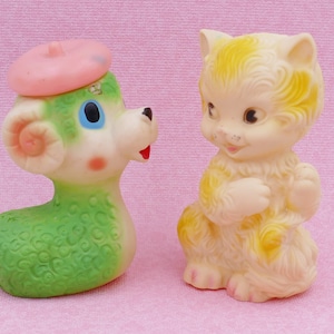 May include: Two vintage rubber toys, a green lamb with a pink beret and a yellow cat, are facing each other on a pink background.