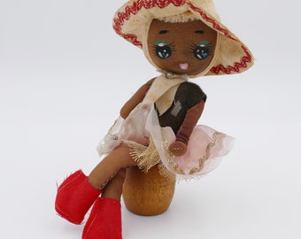 Pode incluir: Uma boneca vintage de pele morena, vestida com um vestido branco e marrom com uma saia de tule rosa, um chapéu de palha marrom com acabamento vermelho e sapatos vermelhos. A boneca está sentada em uma base de madeira.