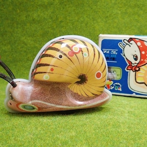 May include: A yellow and brown wind-up snail toy with red and black antennae. The snail is on a green surface and has a white rim around the shell. The box for the toy is in the background and shows a cartoon snail with a red and white polka dot hat.