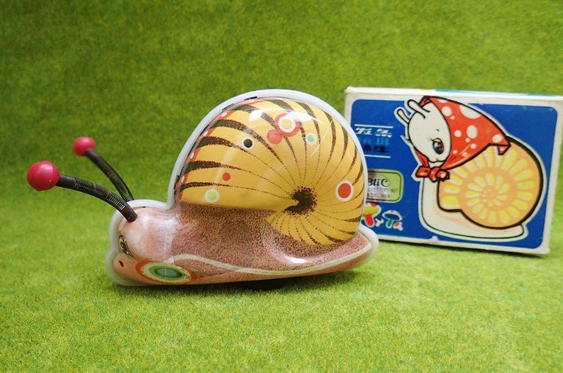 May include: A yellow and brown wind-up snail toy with red and black antennae. The snail is on a green surface and has a white rim around the shell. The box for the toy is in the background and shows a cartoon snail with a red and white polka dot hat.