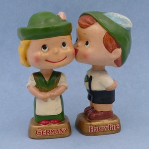 May include: A pair of bobblehead figurines dressed in traditional German clothing. The figurines are kissing and are labeled "Happy Kids" and "Germany".