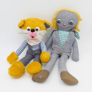 Op de afbeelding: Een gele en witte pluche kattenpop met een blauw-wit geruit overhemd en een blauwe overall zit naast een grijze en blauwe lappop met een blauw-wit geruit overhemd en een blauwe spijkerbroek.