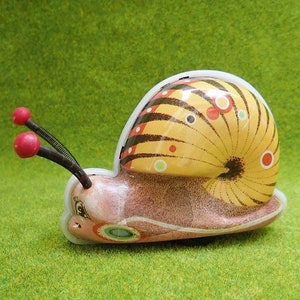 May include: A yellow, brown, and red wind-up snail toy with black antennae and red knobs. The snail has a detailed shell with black stripes and white and red dots.