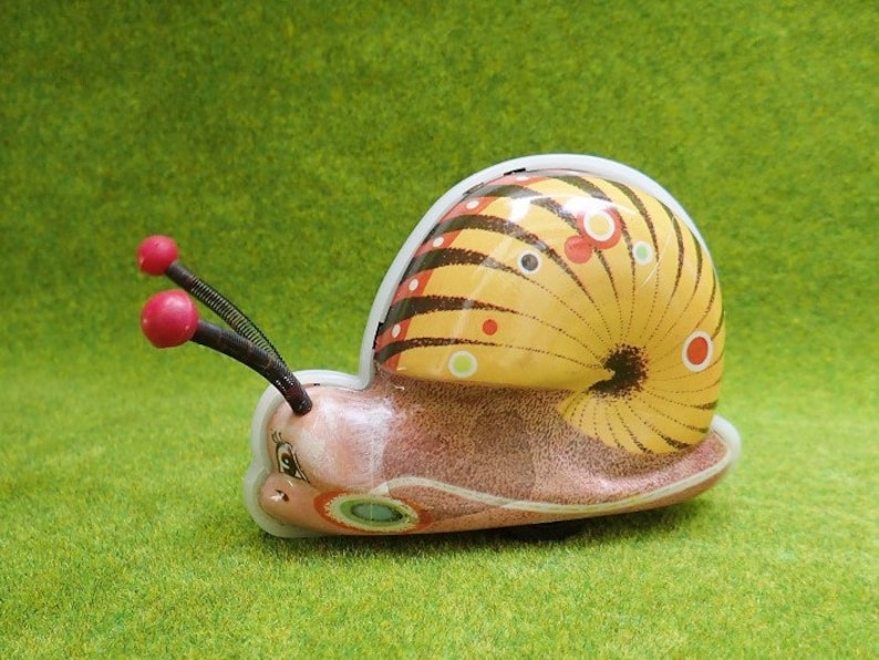 May include: A yellow, brown, and red wind-up snail toy with black antennae and red knobs. The snail has a detailed shell with black stripes and white and red dots.