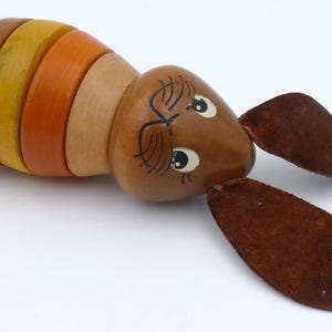May include: A wooden toy shaped like a rabbit with a segmented body in shades of brown, yellow, and orange. The rabbit has painted eyes and long, brown, suede-like ears. The toy is lying on a white surface.