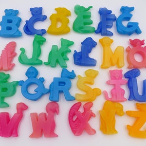 Może przedstawiać: Pełny zestaw alfabetu z kolorowych, przezroczystych plastikowych liter, każda zaprojektowana w kształcie zwierzęcia. Litery ułożone są na białej powierzchni, a każda litera ma inny kolor, w tym niebieski, zielony, żółty, pomarańczowy i różowy.