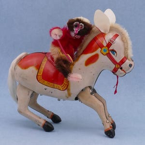 May include: Vintage toy horse with a monkey rider. The horse is painted cream with red accents on the saddle and bridle. The monkey is brown and red, with a pink pom-pom. The toy is set against a light blue background.