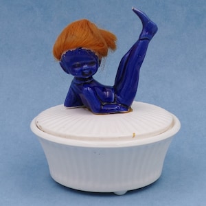 Puede incluir: Una figurita de cerámica azul de una niña con cabello naranja, sentada en una caja de música de cerámica blanca con tapa.