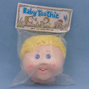 Puede incluir: Una cabeza de muñeca de plástico con cabello amarillo rizado, ojos azules y una boca sonriente. La cabeza está empaquetada en una bolsa de plástico transparente con una etiqueta que dice "Baby Toothie" y "4 1/2" Yellow Curly Hair".