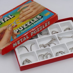 Puede incluir: Una caja roja con las palabras "Merit Metal Puzzles" en la tapa. La caja está abierta y contiene 12 rompecabezas de metal en una bandeja blanca.