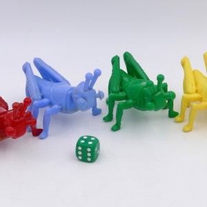 Puede incluir: Cuatro piezas de juego de saltamontes de plástico en rojo, azul, verde y amarillo. Un dado verde con puntos blancos está en el centro de la imagen.