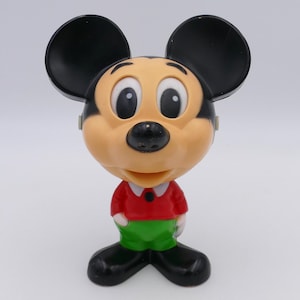 Puede incluir: Una figura de plástico en blanco y negro de Mickey Mouse con una camisa roja y pantalones verdes. La figura tiene grandes orejas negras y una cara sonriente.